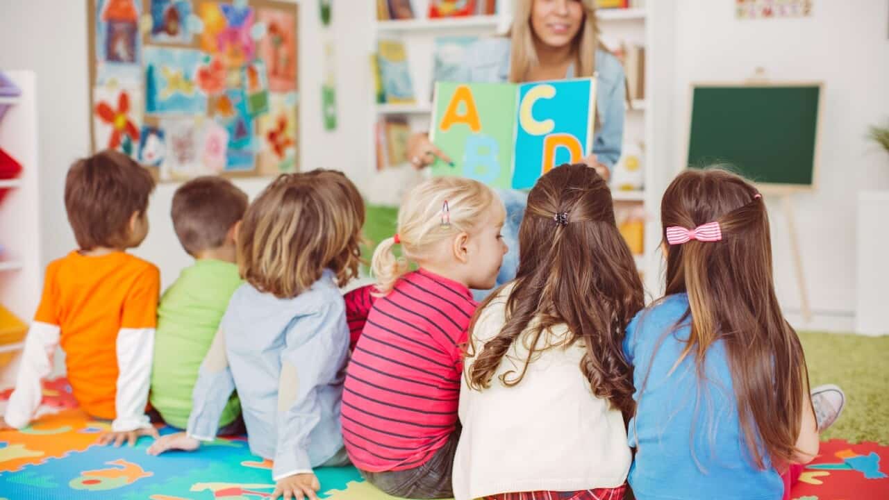 A teacher shows alphabet cards to a group of young children sitting on a colorful play mat in a classroom.