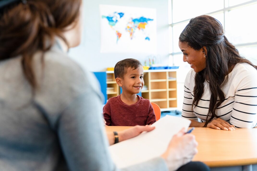 A young boy sits smiling between two adults at a table in a classroom setting, with a world map visible in the background.