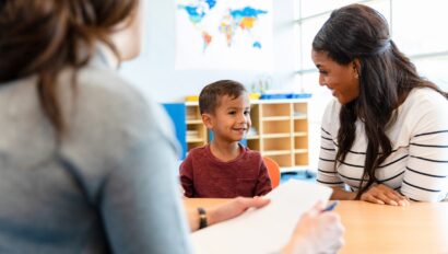 A young boy sits smiling between two adults at a table in a classroom setting, with a world map visible in the background.