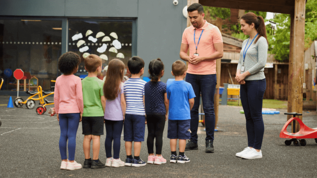 Two adults stand facing a group of young children lined up outside near playground equipment at a daycare or school.